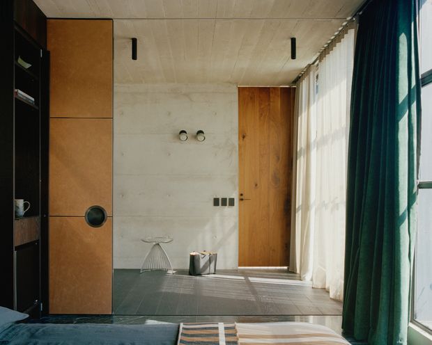 A minimalist bedroom interior with a low bed in the foreground, dark built-in shelving and warm timber cabinetry on the left, a timber door, concrete-look walls, and a full-height glazed opening with sheer and deep green curtains on the right. Natural sunlight creates a calm, textural atmosphere, while a small sculptural chair and decorative objects sit near the door.