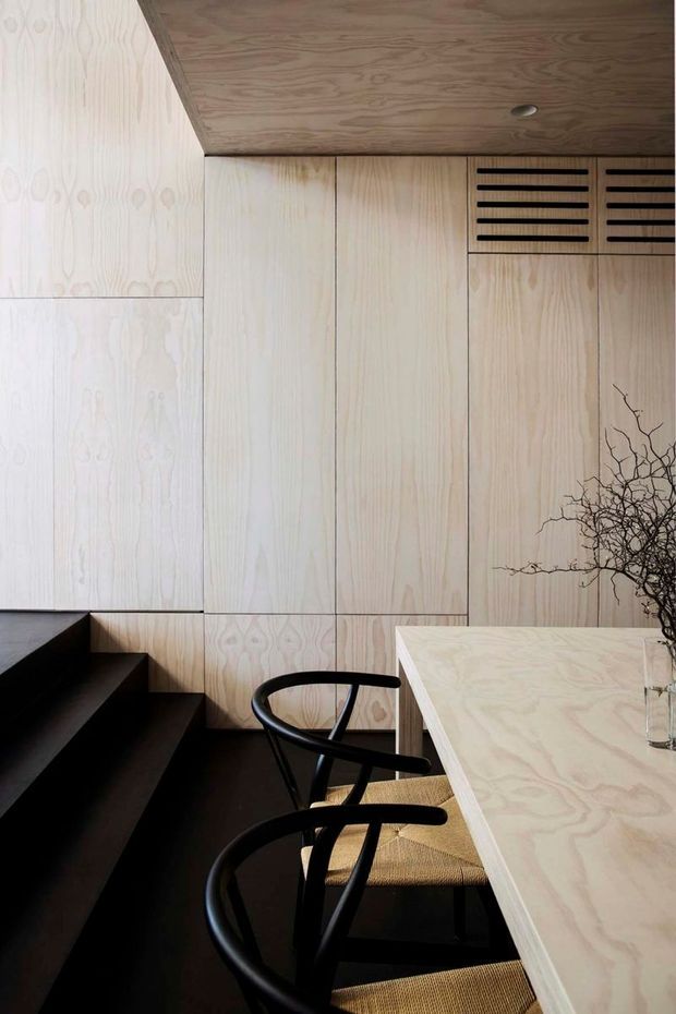 A refined, minimalist interior featuring pale timber wall panelling and cabinetry, a matching timber ceiling, and a pale timber dining counter or table. Black stairs and sculptural black dining chairs create strong contrast, while slatted timber ventilation panels and a small branch arrangement add detail.