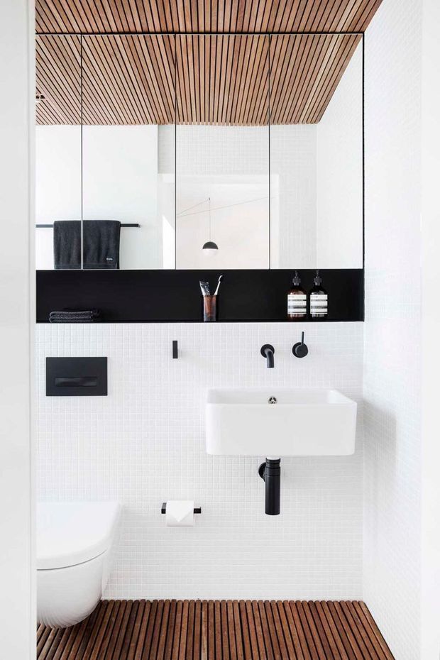 Compact contemporary bathroom with a wall-mounted white basin, black tapware and flush plate, black-framed mirrored cabinetry, and a black open shelf. White small-format tiled walls are paired with a warm timber slatted ceiling and matching slatted floor detail. A wall-hung toilet and folded dark towels are visible.