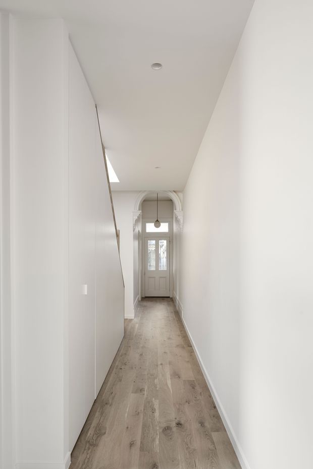 A long, narrow, bright residential hallway or entry passage with white walls, full-height white built-in cabinetry along the left side, pale timber flooring, and a white front door at the far end. The door is framed by ornate arched architectural moulding, with a small pendant light and recessed ceiling lights. The space has a clean, refined, traditional-contemporary appearance.