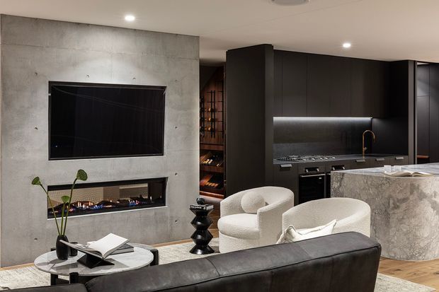 A sophisticated open-plan living room and kitchen with a dark contemporary palette. The room features a raw concrete fireplace wall with built-in linear gas fire and wall-mounted television, charcoal built-in kitchen cabinetry, a stone island with waterfall edge, integrated appliances, and a wine display area. Curved cream lounge chairs and a black leather sofa contrast with the darker architectural finishes.
