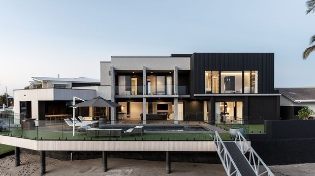 Front exterior view of a large contemporary multi-level waterfront residence with a contrasting rendered concrete and dark vertical-clad facade. The house features broad glazed openings, recessed balconies, a long swimming pool and landscaped terrace, with a raised deck and stair access in the foreground.
