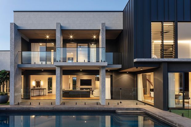 Contemporary multi-level residence viewed from the pool, featuring a flat roof, a mix of pale rendered masonry and dark vertical cladding, exposed concrete columns, expansive glazing, glass balcony balustrades, and warm illuminated interiors.