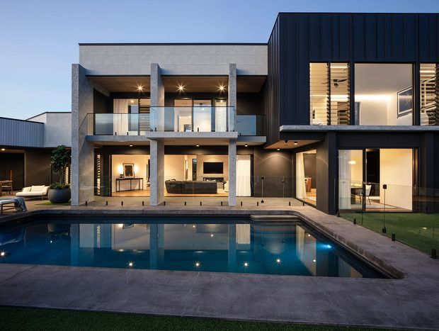 Contemporary two-storey residence viewed from the pool courtyard at dusk. The facade combines dark vertical metal cladding with pale rendered or concrete surfaces, deep overhangs, exposed concrete columns, extensive glazing, and a first-floor glass balustrade. Warm interior lighting is visible through the openings, with a large rectangular swimming pool and paved terrace in the foreground.