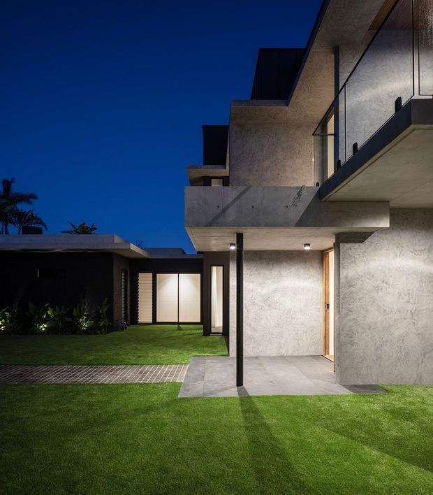 Night-time exterior view of a contemporary multi-level residence organised around a landscaped courtyard. The building features projecting concrete balconies and roof forms, textured grey rendered walls, extensive dark-framed glazing, a slim black structural column, and paved terraces opening onto a manicured lawn. Warm interior lighting contrasts with the deep blue sky and dark landscaping.