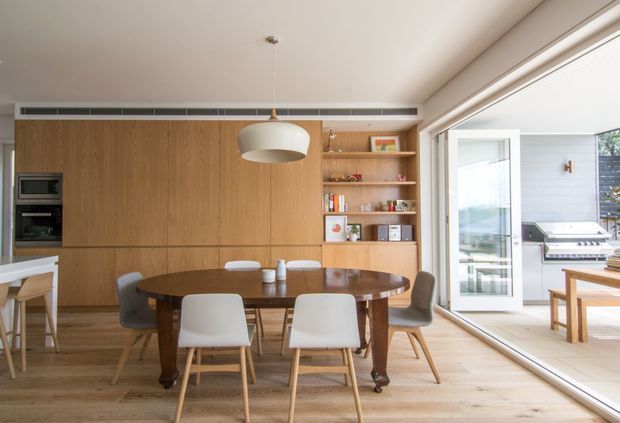 A bright, open-plan dining area with a large oval dark timber dining table and modern upholstered chairs. The room features extensive built-in warm timber cabinetry, integrated appliances, open display shelving, pale timber flooring, and a large glazed opening leading to an outdoor entertaining area with a barbecue. A sculptural pendant light hangs above the table.