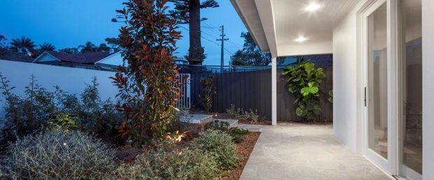 Contemporary covered entry patio beside a modern home, featuring pale textured floor paving, white-framed full-height glazing and doors, recessed ceiling lights, dark vertical privacy fencing, and a landscaped garden bed with mature planting. The view is captured at dusk, with the illuminated entry creating a calm, welcoming atmosphere.