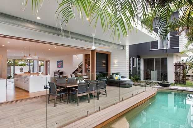 Modern indoor-outdoor living area opening onto a timber deck and swimming pool. The image shows a contemporary kitchen and dining space with a large island, glass dining table, built-in shelving, and lounge seating, connected through full-height frameless glazing to the pool terrace. Palm planting and a neighbouring modern facade are visible outside.