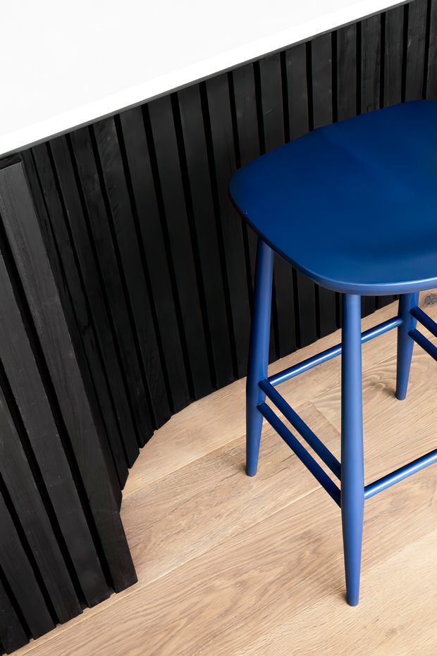 Close-up interior detail showing a vivid blue bar stool with a rounded seat and tubular legs on a light timber floor. Behind it is a dark vertical timber slat wall with a white upper wall or ceiling edge. The image is tightly framed around the furniture and finishes, with no broader room layout visible.