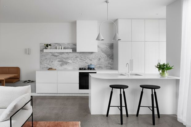 A bright, minimalist open-plan kitchen with white flat-panel cabinetry, a large white island with integrated sink, black bar stools, and sculptural pendant lights. A grey veined stone splashback contrasts with the predominantly white interior. The space opens toward a living area with a sofa and built-in bench seating, while sheer curtains provide soft daylight.