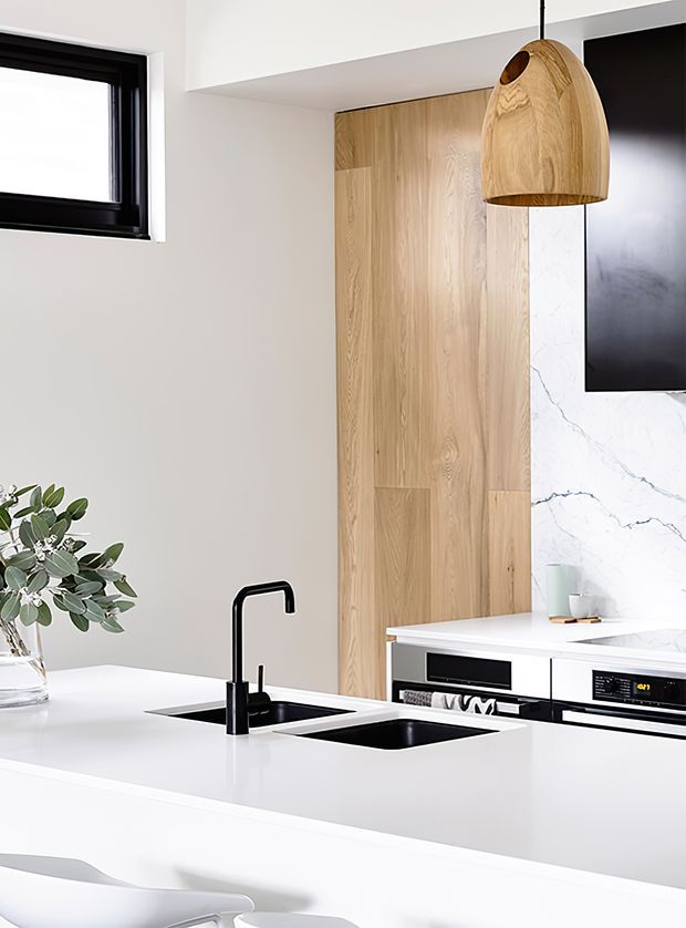Bright contemporary kitchen featuring a large white island with integrated black sink and matte-black gooseneck tap, white cabinetry and benchtops, a vertical light timber wall panel, and a white marble-look splashback. Black-framed window, dark appliances, and a sculptural timber pendant provide contrast.