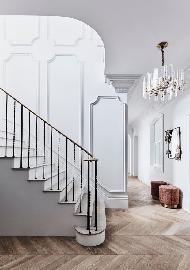 A bright, refined residential hallway and stairway with an elegant curved staircase, black metal balustrade, and brass-toned handrail. White decorative wall panelling, ornate cornices, arched openings, a crystal chandelier, and chevron-patterned timber flooring create a sophisticated classical interior. Two blush upholstered stools and a decorative wall mirror are visible along the corridor.