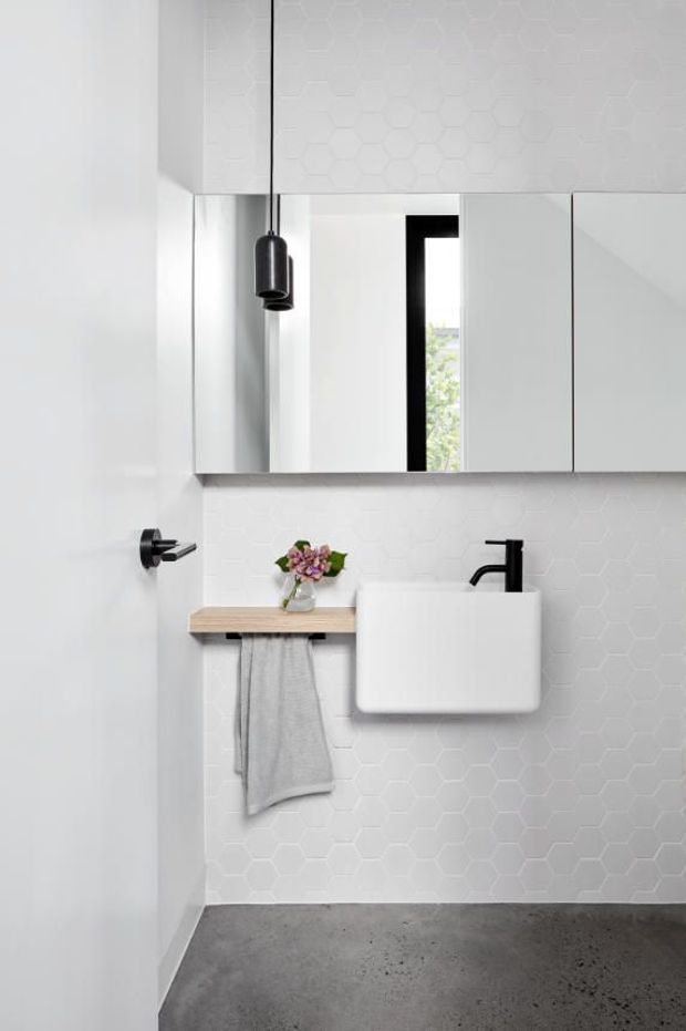 Compact minimalist bathroom with a wall-mounted white basin, black tapware and pendant light, mirrored overhead cabinetry, white hexagonal wall tiles, a light timber shelf with a hanging towel, and a grey concrete-look floor. The room is bright and predominantly monochromatic with subtle timber and black accents.