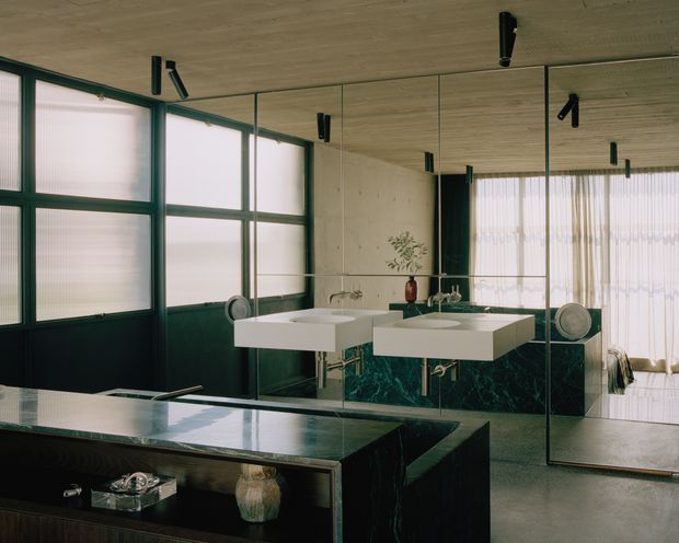 A sophisticated contemporary bathroom featuring twin floating white vessel-style basins on a dark green marble vanity, extensive mirrored walls, dark green-framed ribbed glass windows, polished concrete surfaces, and a wood-look ceiling. Black track lighting and warm sheer curtains create a refined, moody atmosphere.