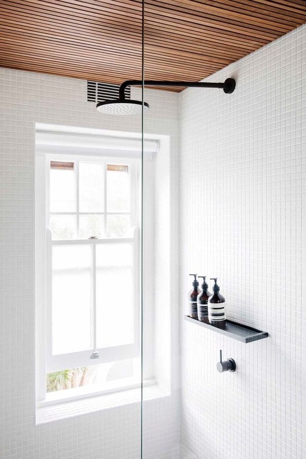 Bright minimalist shower enclosure with white small-format tiled walls, a frameless glass screen, matte black shower hardware and recessed shelf, and a warm slatted timber ceiling. A large white-trimmed window provides abundant natural light.
