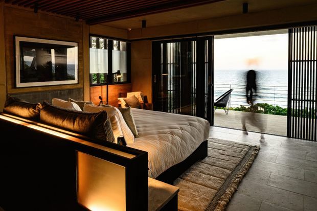 A warm, dark-toned bedroom with a low black bed, textured neutral bedding, and a large woven rug. Timber wall panelling and a slatted timber ceiling create a rich architectural backdrop, while full-height black-framed glazed doors with vertical screening open to a bright waterfront-facing terrace. Integrated amber lighting and carefully framed artwork add a refined resort-like atmosphere.