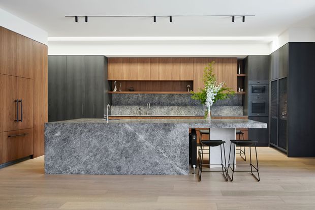 A contemporary kitchen with a large waterfall island clad in dramatic grey stone, integrated seating for two, dark charcoal cabinetry, and warm timber overhead cabinetry and full-height storage. The kitchen includes a textured stone splashback, concealed appliances, timber flooring, and minimalist black track lighting. A vase of foliage provides a natural accent.