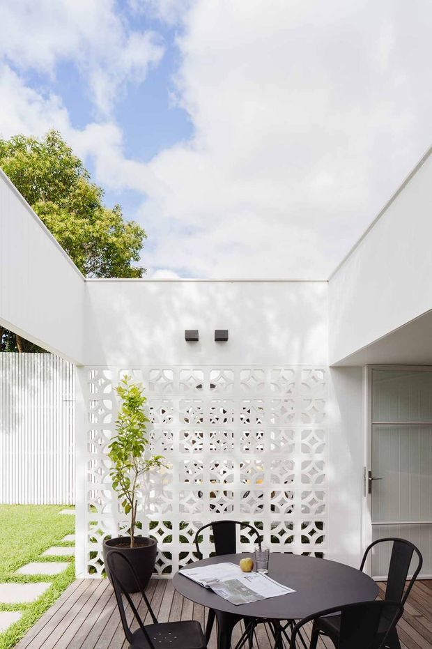 A bright, open-air courtyard dining area with a white rendered architectural enclosure, patterned breeze-block privacy screen, timber decking, and a small black outdoor dining table with chairs. A glazed or screened opening and door are visible on the right, while lawn, stepping stones, and established greenery extend beyond the courtyard. The composition has a clean contemporary feel with strong geometric forms and abundant natural light.