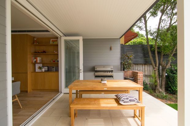 Covered outdoor entertaining area with a timber dining table and bench seating, built-in stainless-steel barbecue, tiled floor, and a light grey weatherboard wall. Large glazed sliding doors open into an interior living or dining space with timber built-in shelving. A timber fence, brick barbecue surround, and established garden are visible beyond.