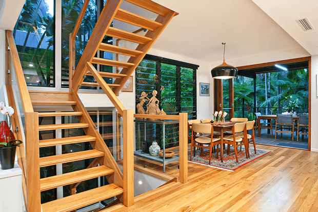 Bright contemporary open-plan dining and stair hall with extensive glazing overlooking lush greenery. The room features a sculptural open-riser timber staircase with timber balustrade, warm timber flooring, a timber dining table with upholstered chairs, a large black pendant light, and black-framed windows and doors. A lower-level void and additional dining or living area are visible beyond the glass balustrade.