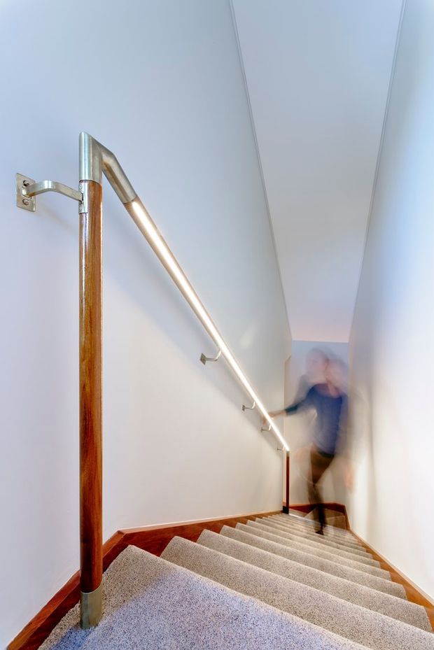A narrow interior staircase descending between white walls, with light carpeted steps, timber skirting and polished timber handrails supported by exposed metal brackets. A slim linear light runs along the wall beside the stairs, creating a clean, contemporary circulation space. A motion-blurred person is visible near the bottom.