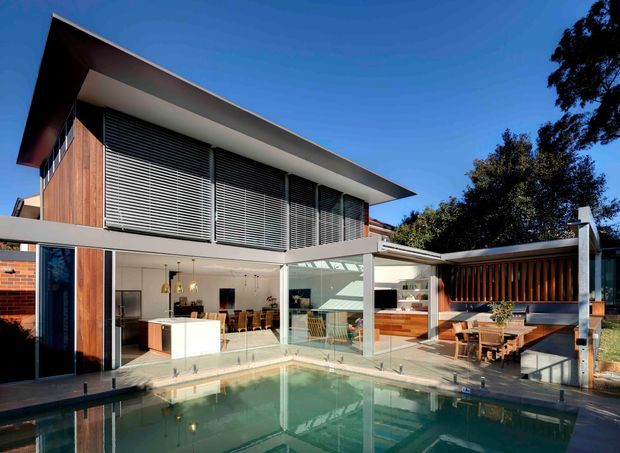 Exterior view of a modern two-storey residence arranged around a swimming pool and outdoor entertaining area. The house features broad floor-to-ceiling glazing, timber wall cladding, extensive horizontal louvres, a dramatic low-pitched roof overhang, and an open-plan kitchen and dining area visible through the glass. A timber-lined outdoor lounge and dining zone sits beside the pool.