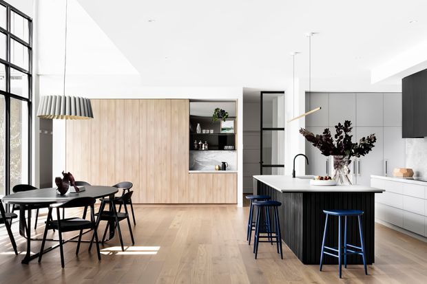 Bright open-plan contemporary kitchen and dining area with pale timber flooring, a large white island with dark vertical-panel cabinetry, integrated white cabinetry, and a light timber feature wall with open shelving. Black-framed floor-to-ceiling glazing, pendant lighting, and dark dining and bar stools add contrast.