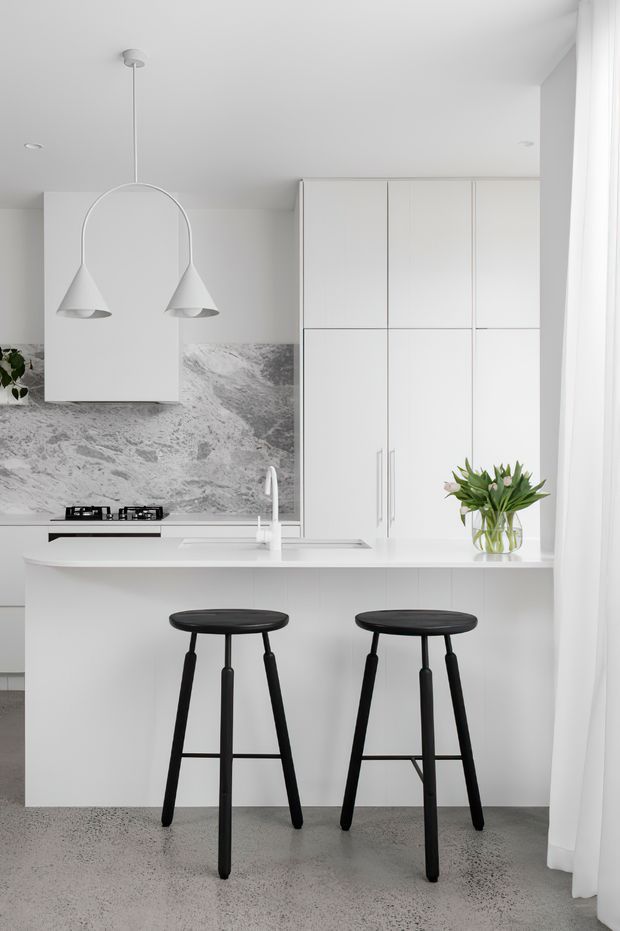 Bright minimalist white kitchen with a compact island and two black bar stools. The space features full-height white cabinetry, a white island benchtop, grey veined stone splashback, integrated cooktop, chrome tapware, pendant lighting, sheer curtains and a polished grey terrazzo-like floor.