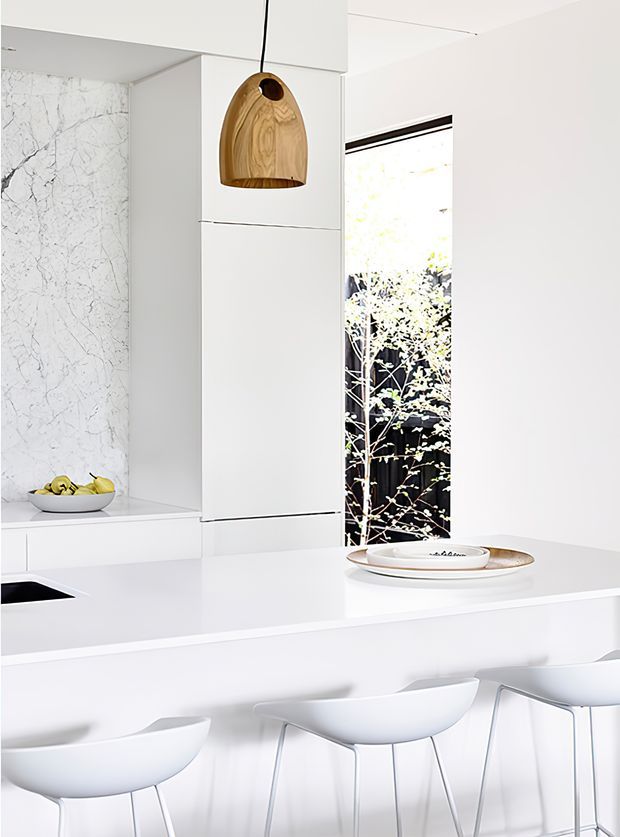 Bright, minimalist white kitchen with a large island and three white bar stools. The kitchen features seamless white cabinetry, a veined marble-look wall surface, a white benchtop, a sculptural timber pendant light, and a narrow window overlooking foliage.