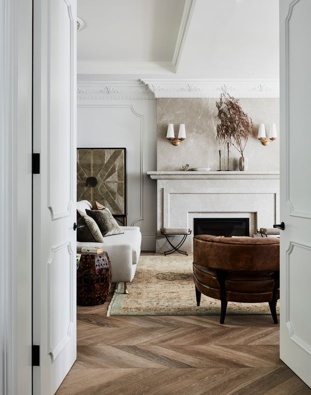 A refined living room viewed through open paneled double doors. The room features ornate wall mouldings and ceiling cornices, a sculpted stone fireplace surround, brass wall sconces, a white sofa, a brown leather lounge chair, an upholstered vintage-style rug, and herringbone timber flooring. The palette is predominantly soft white, beige, taupe, and warm brown, creating an elegant, classic atmosphere.