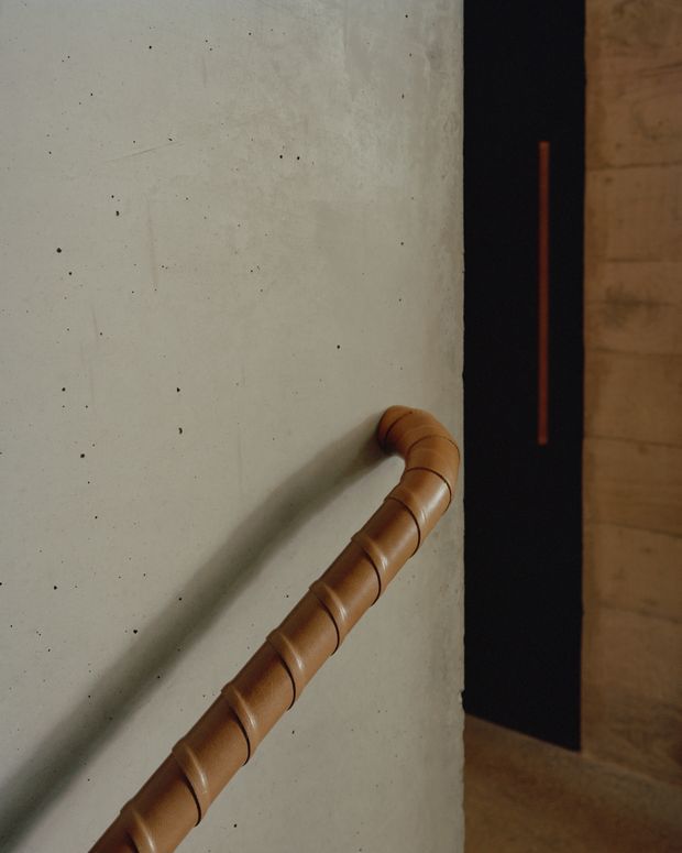 Close-up architectural detail of a warm brown cylindrical timber handrail with segmented carved joints, mounted diagonally against a pale grey textured concrete wall. A dark vertical panel and warm timber wall surface are visible in the background, creating a restrained, tactile contemporary interior composition.