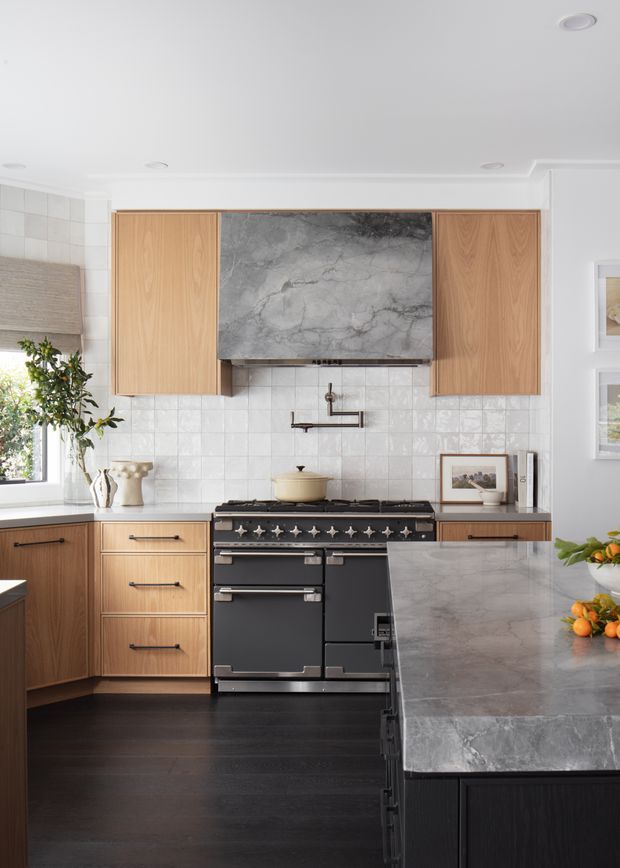 Bright, refined kitchen featuring light timber cabinetry, a large dark veined stone rangehood surround, white square tiled splashback, black range cooker, dark timber flooring, and a substantial grey stone island with dark cabinetry. Natural light enters from the window at left, with framed artwork and restrained styling details.