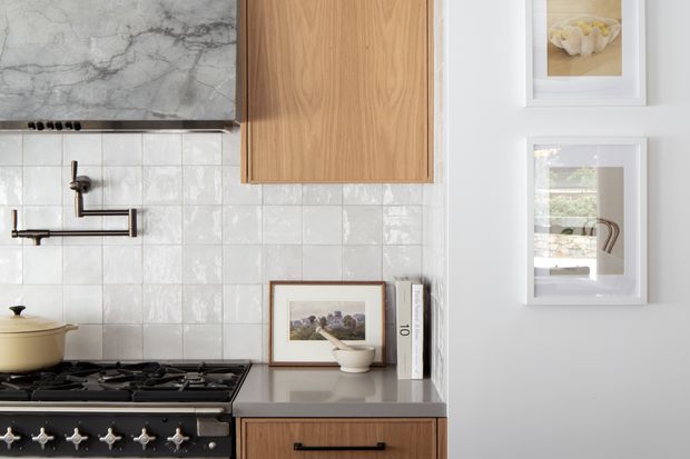 Close-up view of a refined kitchen cooking area featuring a dark stone benchtop, white glossy square wall tiles, timber cabinetry, a gas cooktop with brass-toned controls, and a wall-mounted pot filler. Framed artwork and styling objects add a warm, curated character.