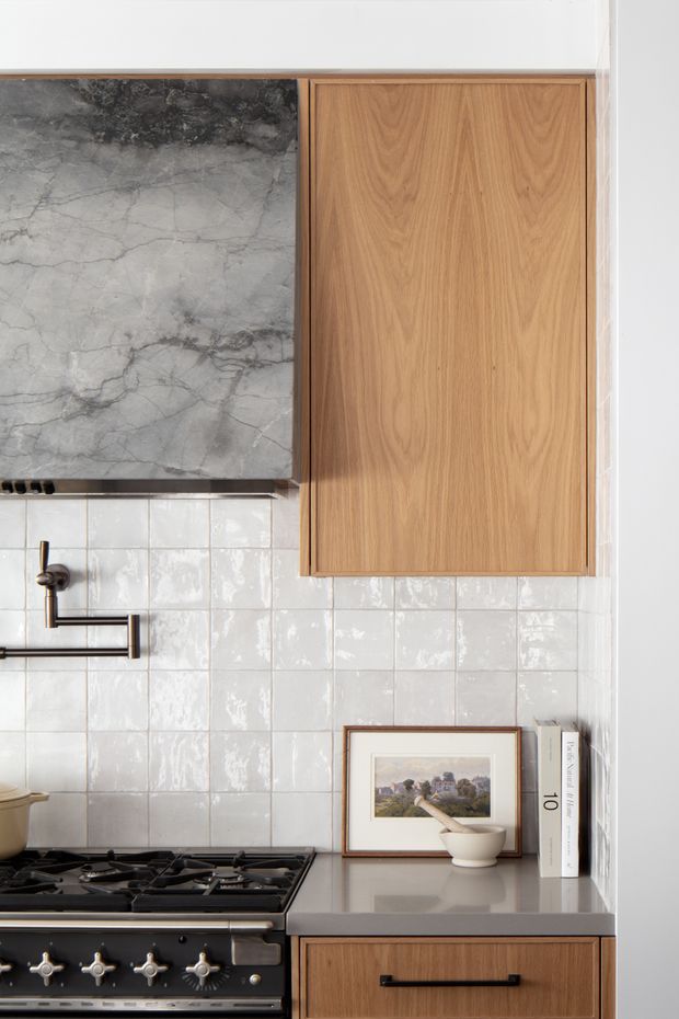 Close-up of a refined kitchen cooking zone featuring a dark veined stone splashback or rangehood surround, glossy off-white square wall tiles, warm natural timber cabinetry, and a grey stone benchtop. A black range cooker, dark metal tap, framed artwork, and styling accessories add contrast. The composition highlights integrated joinery and material detailing in a compact, softly lit kitchen.