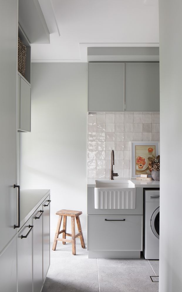 A compact, light-filled utility kitchen/laundry space with pale grey cabinetry, a white ceramic farmhouse sink, glossy square tiled splashback, black hardware, and a front-loading washing machine. A wooden stool and framed artwork add warmth to the otherwise restrained, contemporary interior.