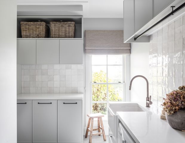 A bright, compact contemporary laundry room with pale grey built-in cabinetry, open shelving with wicker baskets, glossy white square-tile walls, and a long white countertop containing a deep farmhouse-style sink. A tall window with a woven Roman blind provides natural light, while a brushed metal gooseneck tap, black hardware, timber stool, and dried floral arrangement add detail.