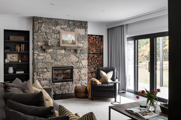 A warm, refined living room with a large rough-stone fireplace wall and integrated firewood storage. Dark upholstered sofas and an armchair surround a low coffee table, while full-height glazed doors with grey curtains open toward an outdoor area. Built-in dark shelving, layered textiles, framed art, and natural firewood add character.