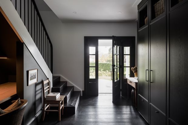 A dark, refined residential entry hall with black-stained timber flooring, charcoal walls, a black-framed glazed front door, a compact staircase with black balustrade, and extensive full-height built-in cabinetry. A timber-lined opening and seating area are visible to the left, with daylight entering through the open door.
