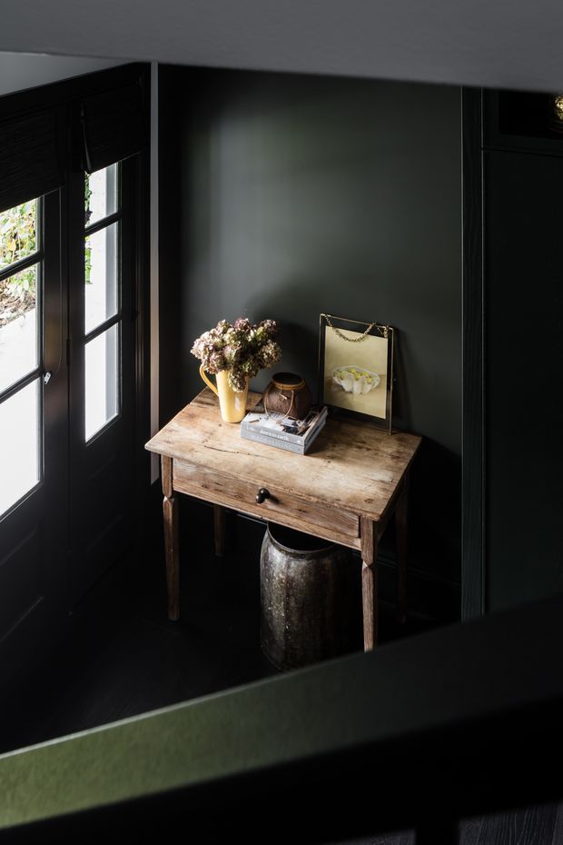 A moody, narrow entryway or hallway corner with black-painted walls, dark timber-framed glazed doors, and dark flooring. A rustic timber console table is styled with a vase of dried flowers, books, a framed artwork, and a metal vessel beneath. The scene is viewed partly through a dark balustrade or railing, creating a layered architectural composition with strong contrast and subdued natural light.