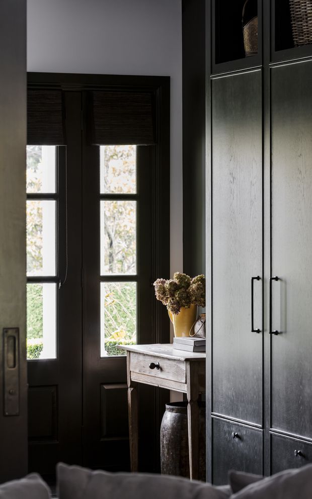 Moody interior vignette featuring a dark-painted glazed entry door with internal blinds, a weathered timber console table, and tall dark built-in cabinetry with inset panels and black hardware. Decorative objects and a vase of dried flowers add warmth, while a sofa edge is visible in the foreground.