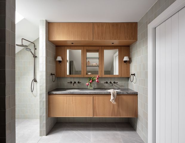 A contemporary bathroom with a floating double vanity in warm timber cabinetry, dark stone benchtop, mirrored wall cabinets, wall-mounted taps, and twin basins. Pale grey-green tiled walls and matching floor surround a walk-in shower with exposed chrome fittings. White paneled doors and simple wall sconces complete the clean, refined space.