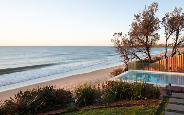 A coastal residential outdoor scene overlooking a sandy beach and calm ocean, with a swimming pool in the foreground, timber privacy fencing, native-style landscaped planting, and a paved garden path. The image is captured from the property, with the beachfront setting dominating the view.