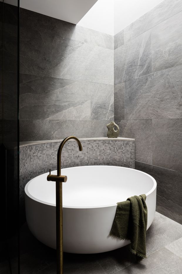 A refined contemporary bathroom featuring a freestanding white oval bathtub, dark bronze floor-mounted bath filler, large-format grey stone-look wall and floor tiles, and a grey mosaic-tiled ledge. Natural light enters from a high skylight or clerestory opening, creating a dramatic yet calm atmosphere.