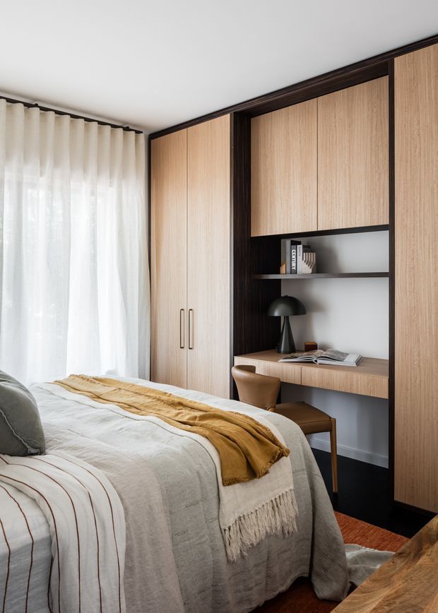 A warm, refined bedroom featuring a built-in timber wardrobe and integrated study nook with open shelving, desk, chair, and black dome lamp. The bed is dressed in soft neutral linen with a mustard throw, beside full-height sheer curtains and a bright window. Dark timber framing and flooring provide contrast against the light oak joinery and white walls.