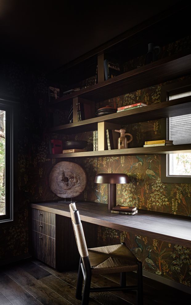 A dark, richly detailed home office with built-in dark timber shelving and a long integrated desk. The shelving displays books, ceramics and sculptural objects against a dramatic botanical wallpaper backdrop. A vintage-style desk lamp, upholstered chair and windows with blinds complete the moody study.