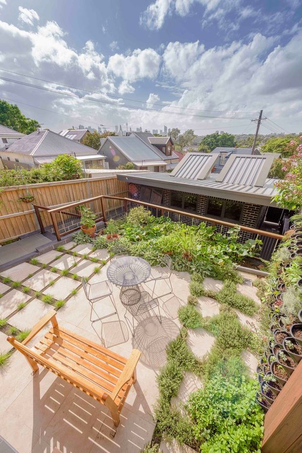 Bright elevated view over a landscaped residential courtyard and patio, with paved stepping stones, garden beds, outdoor wire furniture, timber seating, timber fencing and a low brick-clad house with a distinctive corrugated metal roof. Neighbouring pitched-roof homes and an urban skyline are visible beyond.