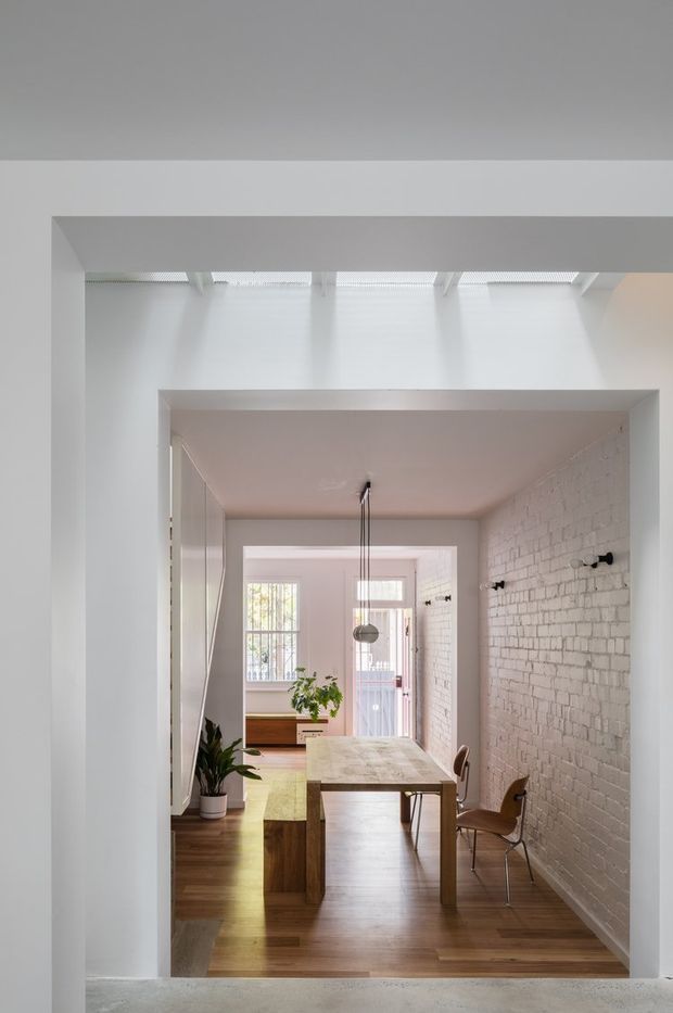 Bright, minimalist dining area viewed through a wide interior opening. The room features warm timber flooring, a light timber dining table with chairs, a white-painted exposed brick wall, simple black wall sconces, potted plants, and abundant daylight entering through high roof glazing and rear windows.