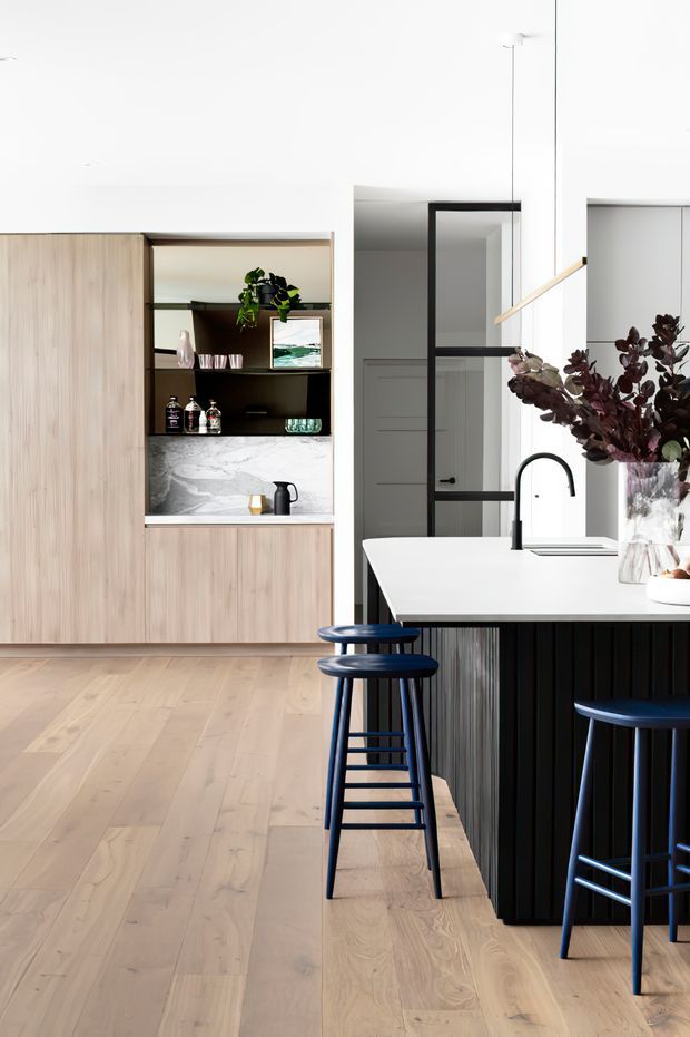 Bright contemporary kitchen interior featuring pale timber cabinetry and wide-plank timber flooring, a white stone island benchtop with a black fluted base, black-framed glass partition doors, and dark blue bar stools. Open shelving displays glassware and decor, while a marble-look splashback and black gooseneck tap add contrast. Minimal pendant lighting and abundant white surfaces create a clean, refined atmosphere.