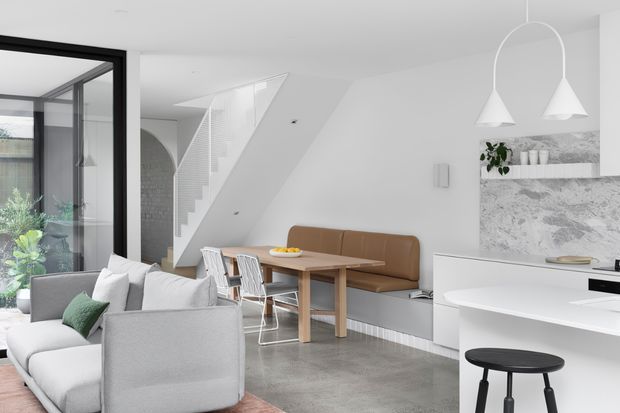 Bright open-plan contemporary interior combining a living area, dining nook, and kitchen. The room features white walls and cabinetry, a large white sofa, a timber dining table with a built-in brown banquette, black-framed glazing, polished grey concrete flooring, and a dramatic grey marble or stone kitchen backsplash. A stairway rises behind the dining area, while sculptural white pendant lights hang over the kitchen island.