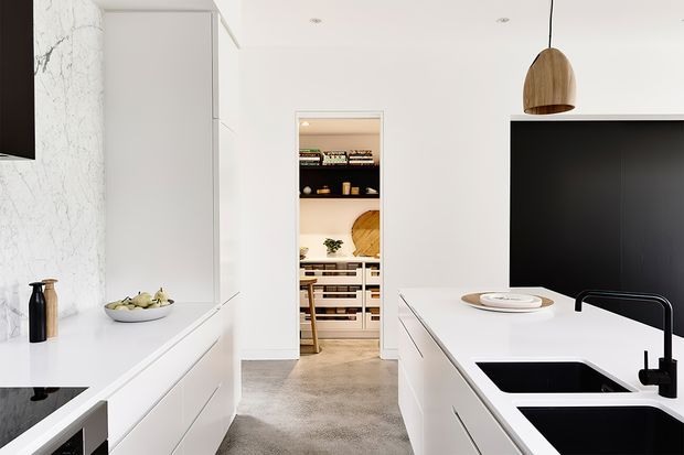 Bright, minimalist white kitchen with a large island, integrated black double sink and black tapware, white cabinetry, a dark stone or black countertop section, and a timber pendant light. A walk-in pantry is visible through the central doorway, with open shelving and organised storage. The room has a pale textured floor and a white marble-look feature wall.